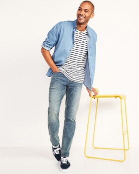 A model leans against a yellow colored stool wearing Athletic Taper style jeans, and a blue denim shirt over a striped top