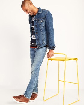 A model leans against a yellow colored stool wearing Slim style jeans, a denim jacket and striped top