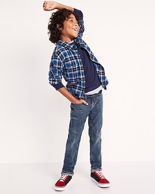 Un jeune garçon porte un jean foncé, des chaussures de sport rouge et bleu marine, un t-shirt et une chemise en flanelle superposée sur le t-shirt. 