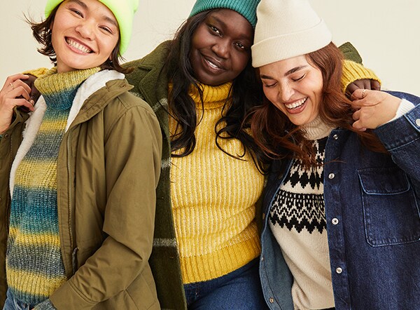 Three female models are wearing turtleneck sweaters, jackets, and beanie hats.