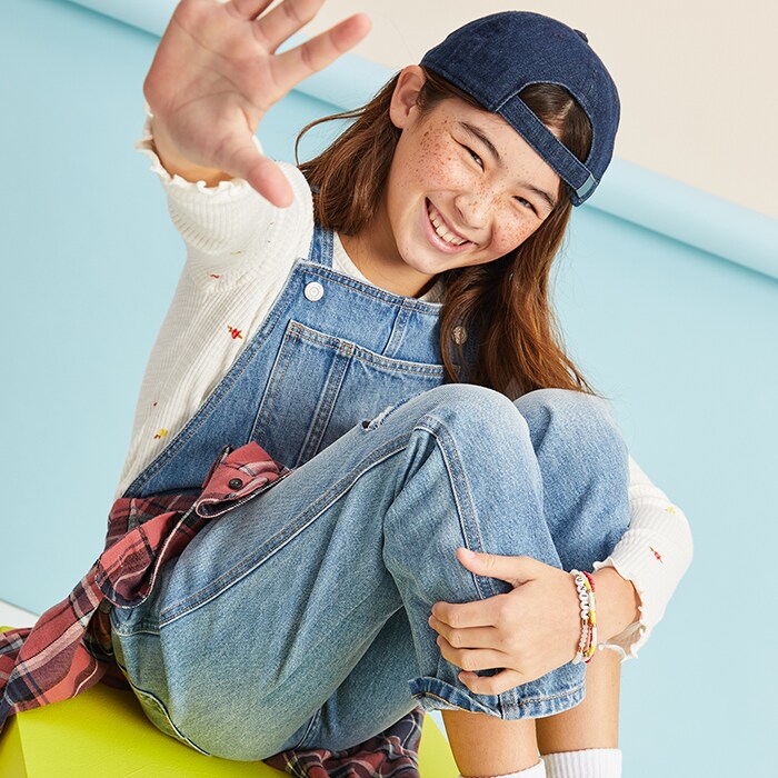A female model wearing jeans overall, white long sleeve shirt and a jean hat.