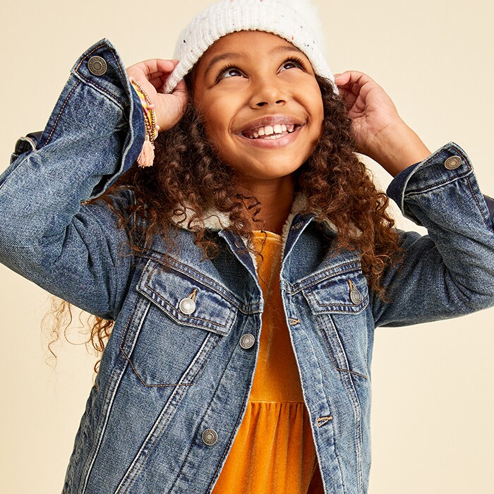 A female model wearing an orange top, jean jacket and a white winter hat.