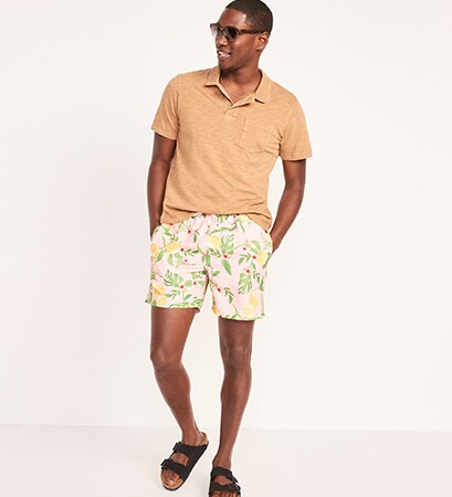 A male model wears a dark cream colored polo shirt & 5 inch floral-printed swim trunks.