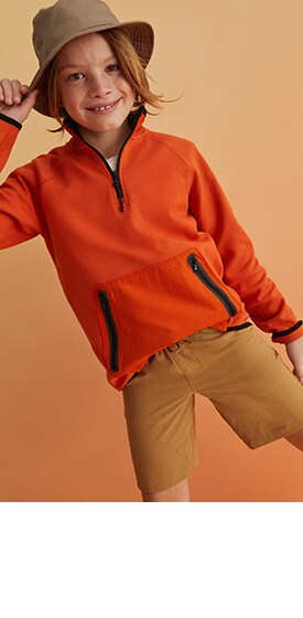 A male model wearing a brown bucket hat, brown shorts, and a bright orange quarter zip.
