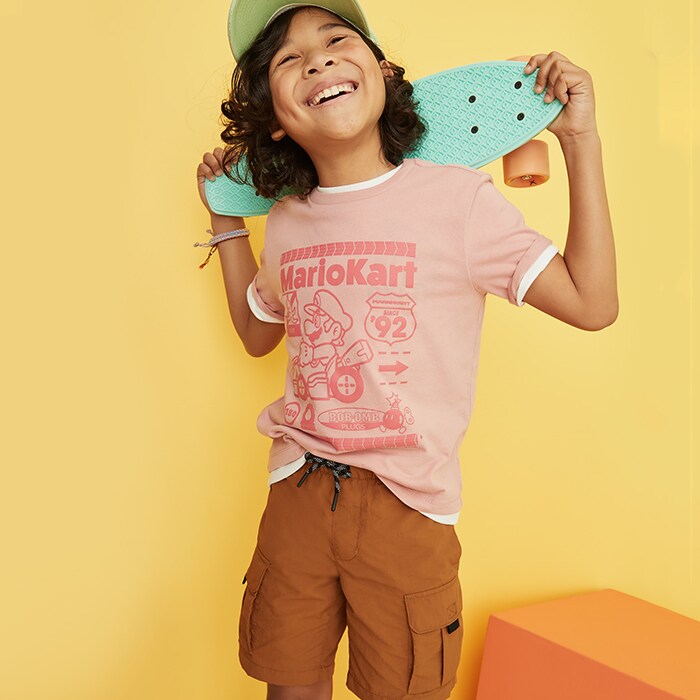 A boy holding a skateboard wearing cargo sorts and a graphic print tee.
