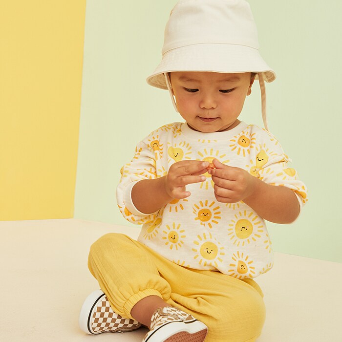 A young male model wearing a yellow bucket hat, yellow checker slip on shoes, yellow pants, and a yellow crewneck with sunshine print.
