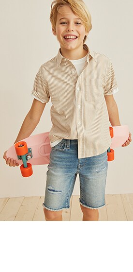 Un garçon porte une chemise à rayures et un short en denim coupé, et il tient une planche à roulettes.