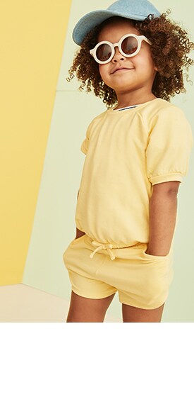 Un tout-petit aux cheveux bouclés porte des lunettes de soleil rondes blanches, une casquette de baseball et une combinaison-short jaune en tissu doux.