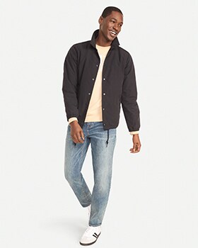 A male model wears Athletic Taper style jeans, and a dark shirt jacket over a cream colored t-shirt.