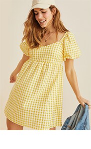 A woman in a checkered yellow and white babydoll style dress, paired with a baseball cap and a jean jacket.