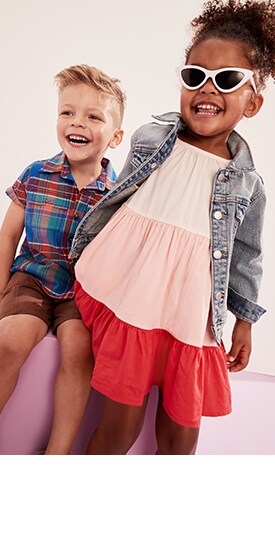Une jeune fille porte une robe trapèze étagée de couleur unie et une veste en denim, tandis qu’un garçon porte une chemise de travailleur à poche et à carreaux en mélange de lin et un short.