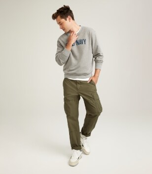 A male model wears cargo style pants & a grey logo crew neck sweatershirt.