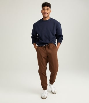 A male model wears brown joggers & a dark crew neck sweatshirt.