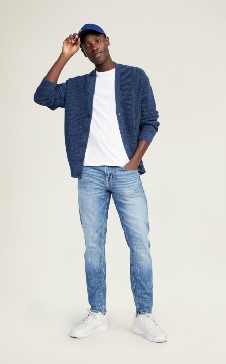 A male model wears light washed Slim Taper style denim & a dark blue cardigan sweater.
